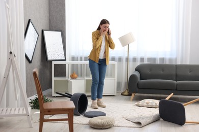 Desperate woman talking on smartphone in messy living room after robbery