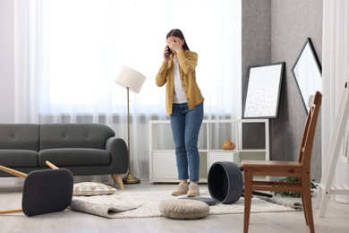 Photo of Desperate woman talking on smartphone in messy living room after robbery