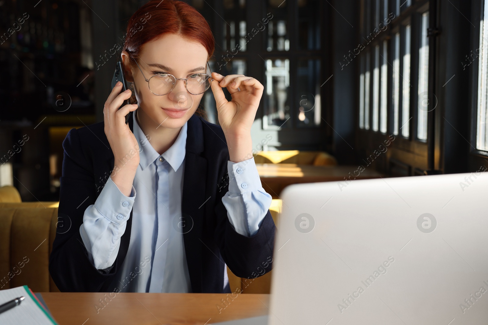 Photo of Young female student with laptop talking on phone while studying at table in cafe