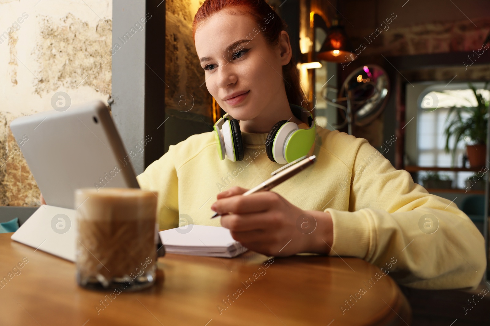 Photo of Young female student with tablet and headphones studying at table in cafe