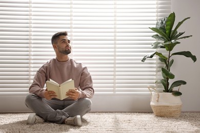 Man reading book near window blinds at home