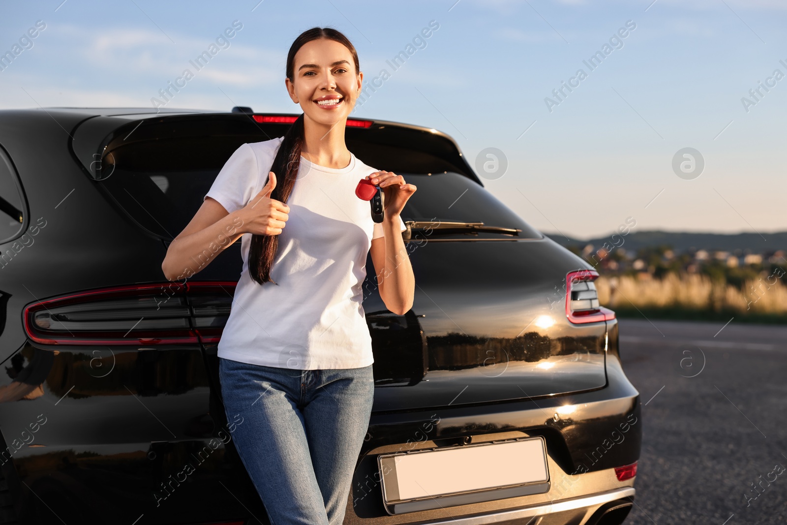 Photo of Happy young woman with key showing thumbs up near her new car outdoors