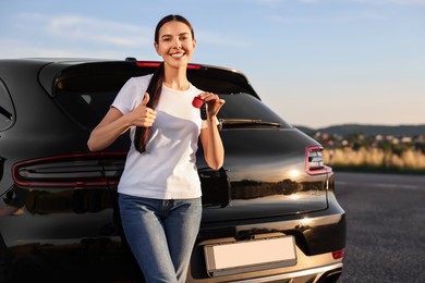 Happy young woman with key showing thumbs up near her new car outdoors