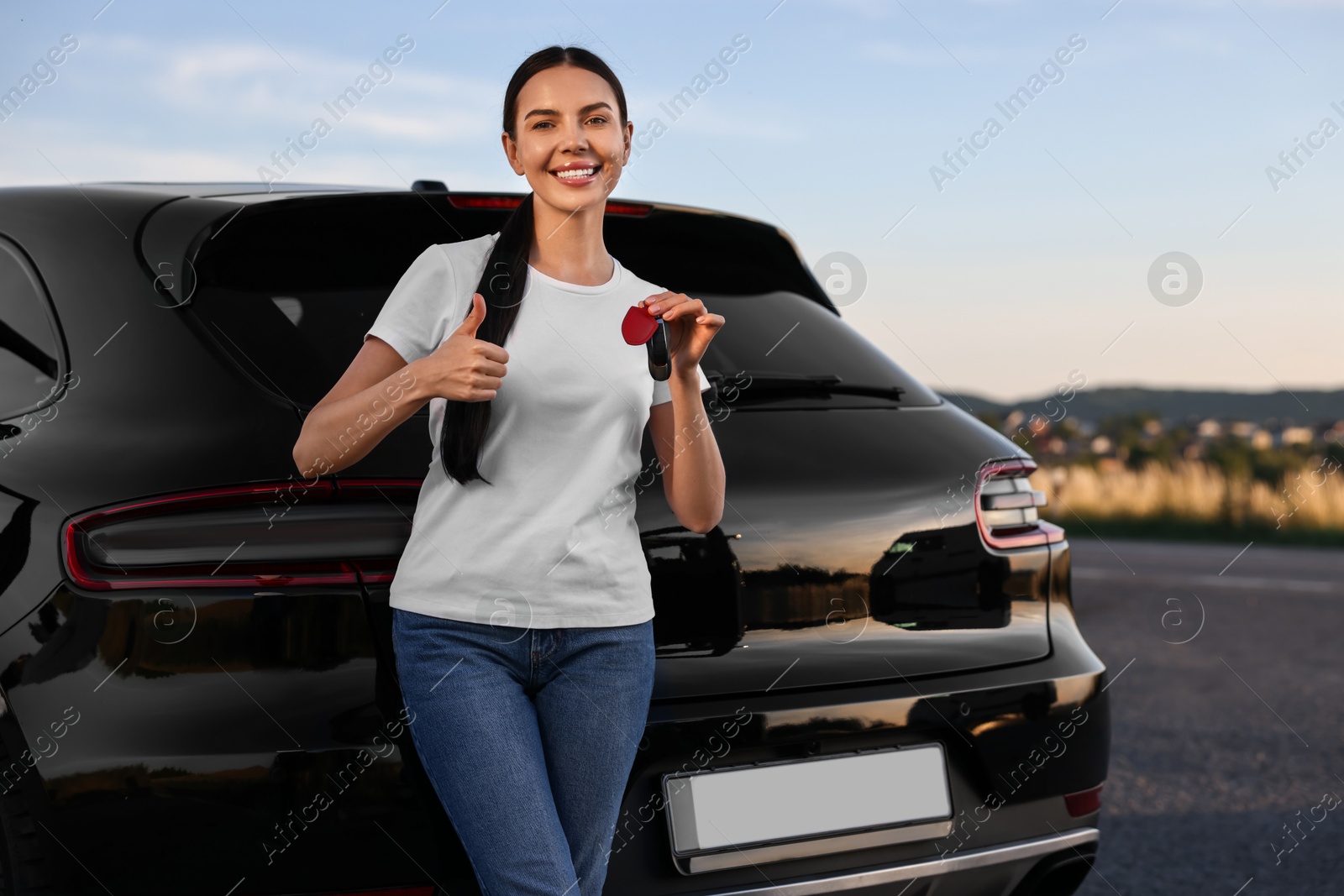 Photo of Happy young woman with key showing thumbs up near her new car outdoors