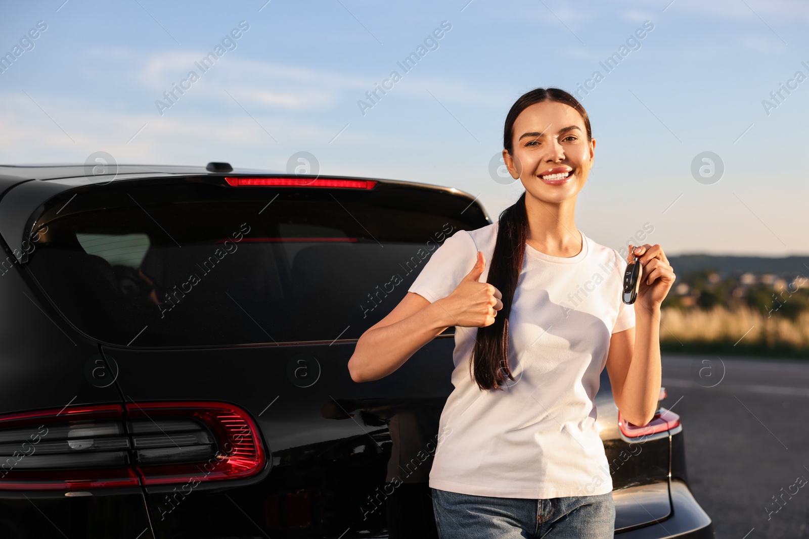 Photo of Happy young woman with key showing thumbs up near her new car outdoors