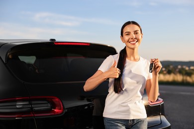 Photo of Happy young woman with key showing thumbs up near her new car outdoors