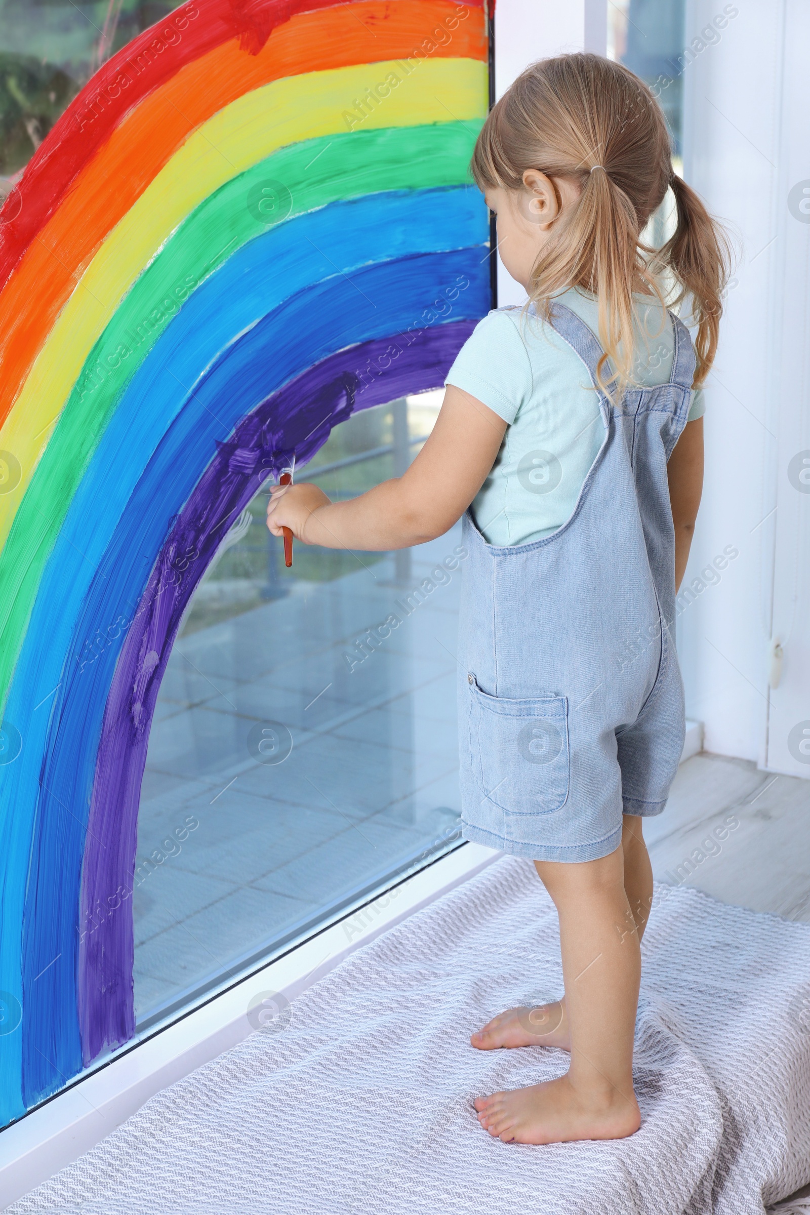 Photo of Little girl drawing rainbow on window indoors