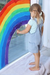 Little girl drawing rainbow on window indoors