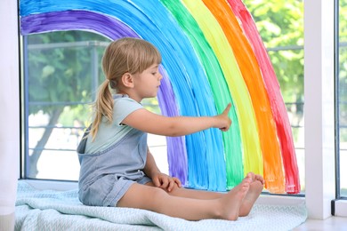 Photo of Little girl touching picture of rainbow on window indoors