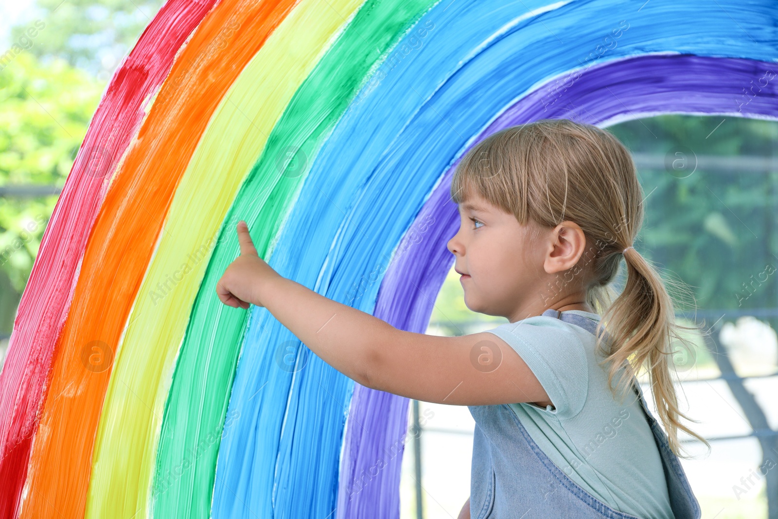 Photo of Little girl touching picture of rainbow on window indoors