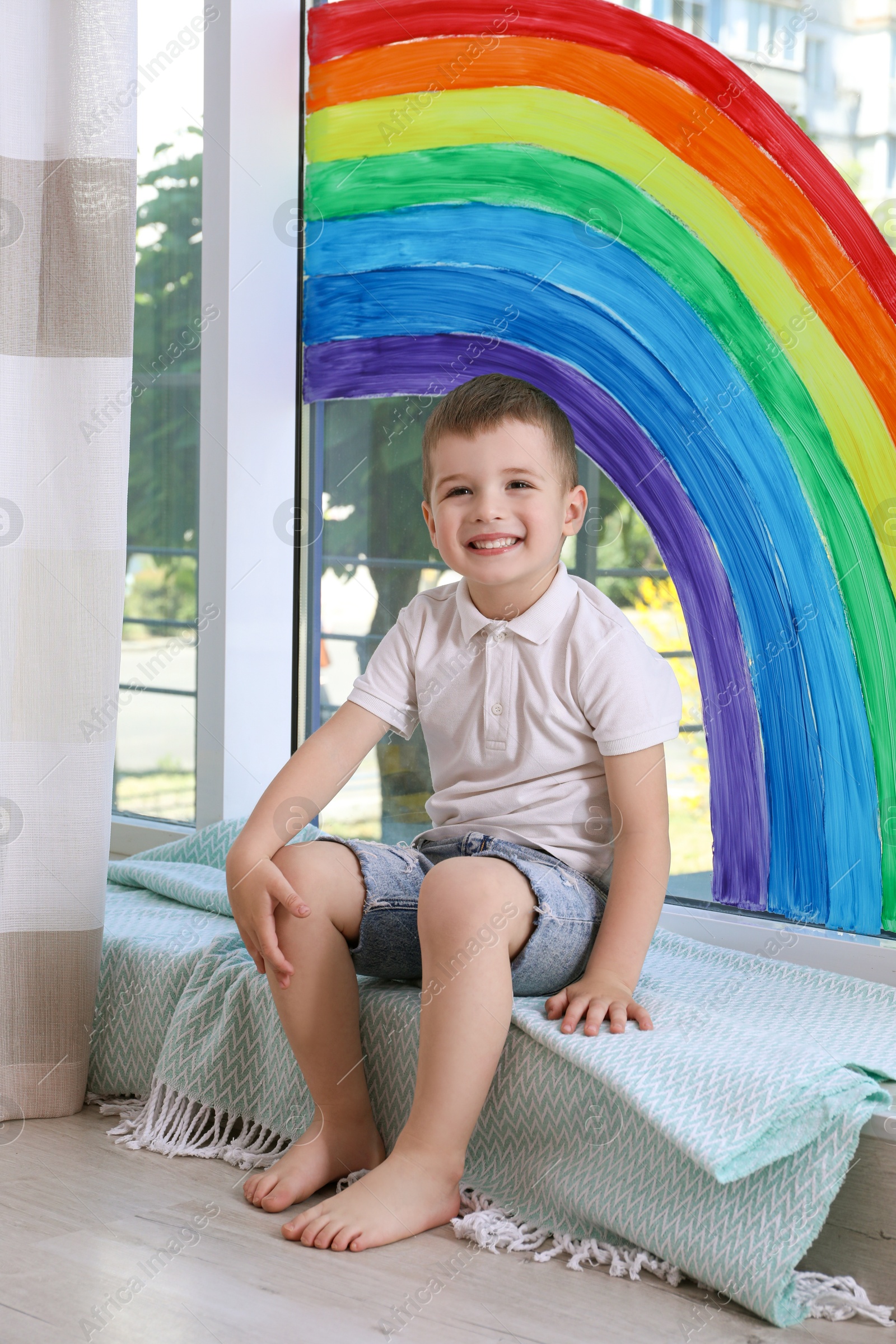 Photo of Little boy near rainbow painting on window indoors