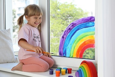 Photo of Little girl drawing rainbow on window indoors