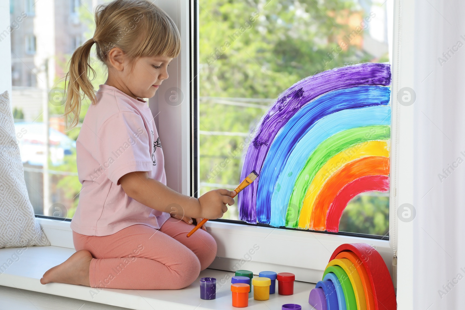 Photo of Little girl drawing rainbow on window indoors