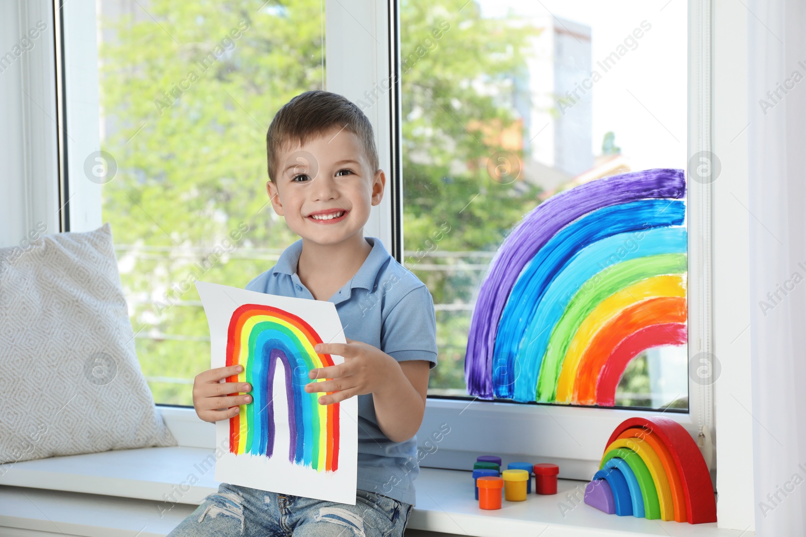 Photo of Happy little boy with picture of rainbow near window indoors