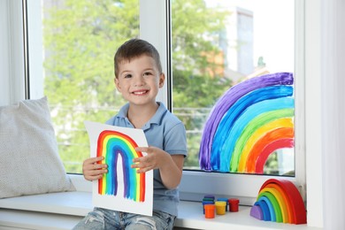 Photo of Happy little boy with picture of rainbow near window indoors