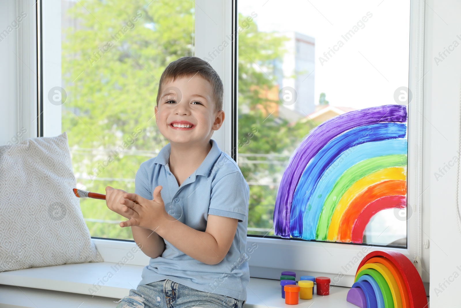 Photo of Little boy with brush near rainbow painting on window indoors
