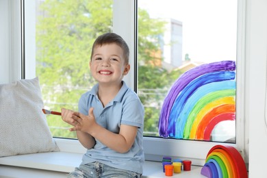 Photo of Little boy with brush near rainbow painting on window indoors