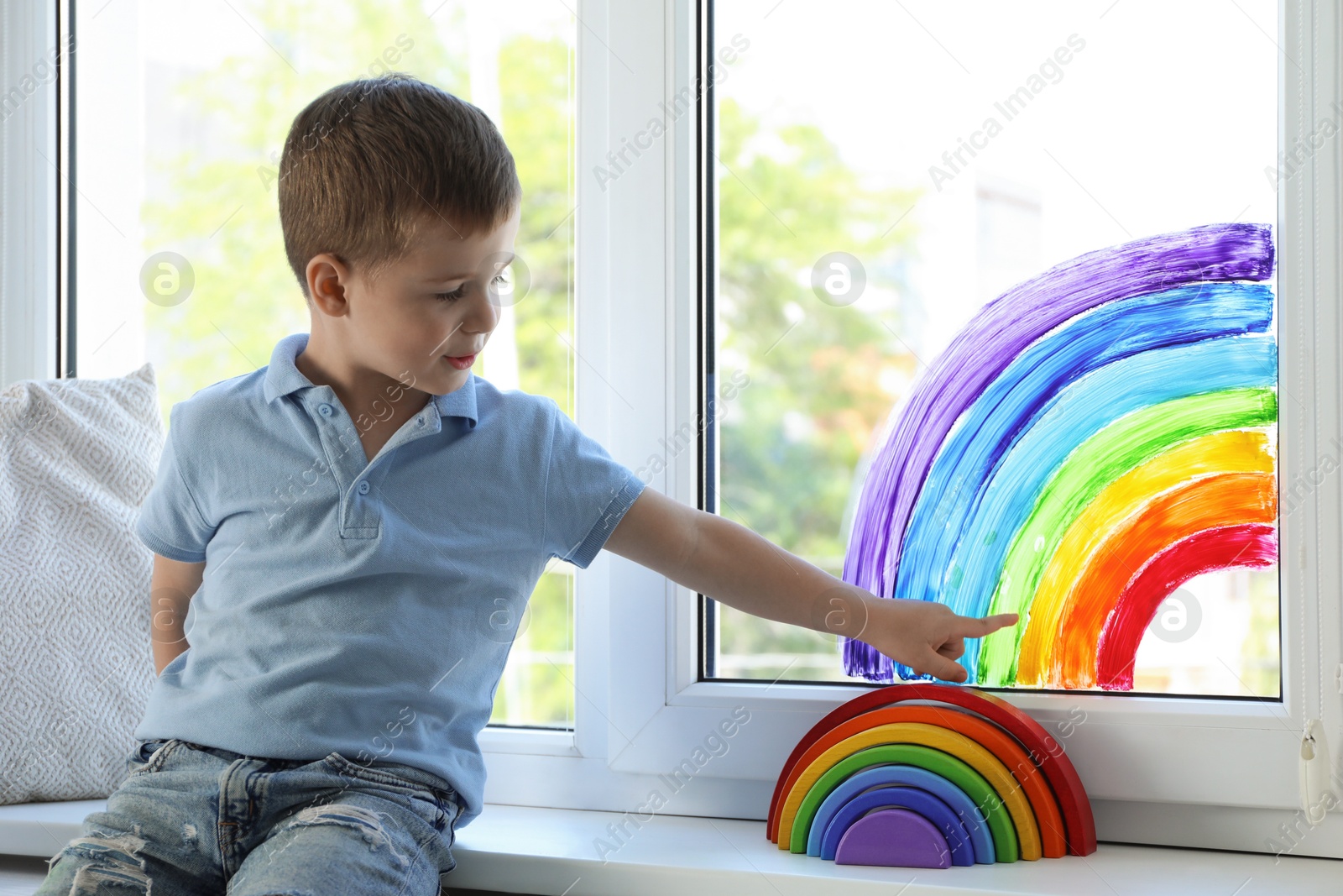 Photo of Little boy touching picture of rainbow on window indoors