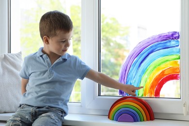 Little boy touching picture of rainbow on window indoors