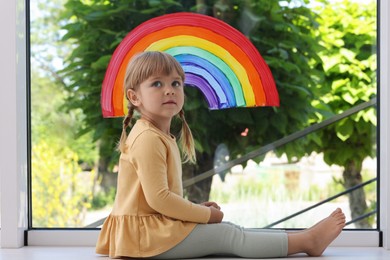 Photo of Little girl near rainbow painting on window indoors