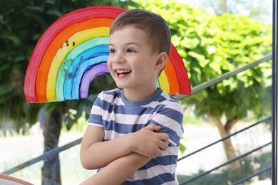 Photo of Happy little boy near rainbow painting on window indoors