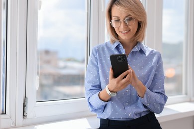 Happy woman using mobile phone near window indoors, space for text
