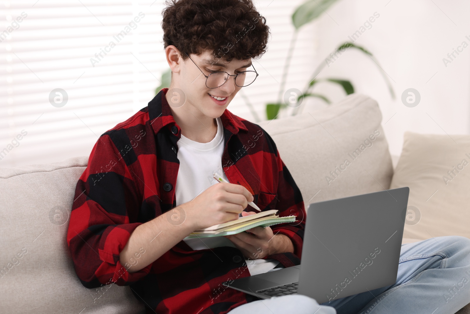 Photo of Teenager taking notes while working with laptop on sofa at home. Remote job