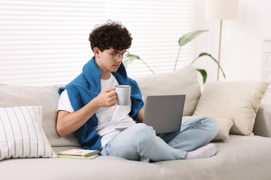 Photo of Teenager holding cup of drink working with laptop on sofa at home. Remote job