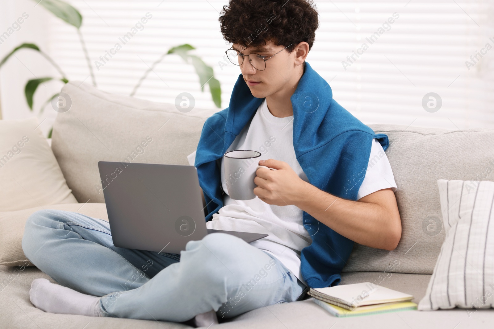 Photo of Teenager holding cup of drink working with laptop on sofa at home. Remote job