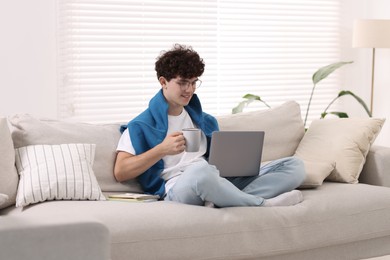 Teenager holding cup of drink working with laptop on sofa at home. Remote job