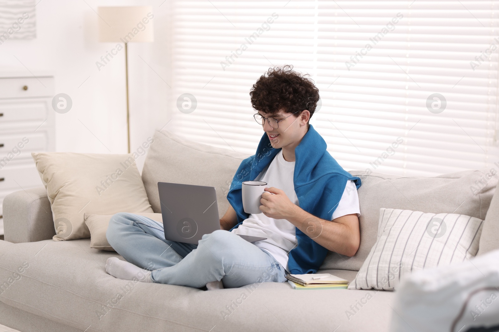 Photo of Teenager holding cup of drink working with laptop on sofa at home. Remote job