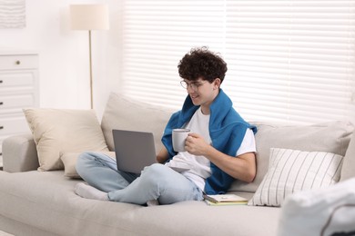 Teenager holding cup of drink working with laptop on sofa at home. Remote job