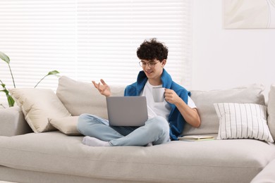 Teenager holding cup of drink working with laptop on sofa at home. Remote job