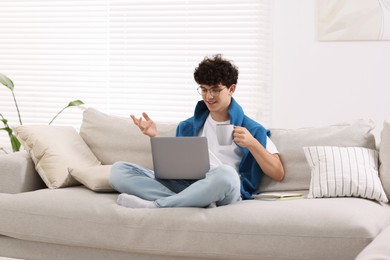 Photo of Teenager holding cup of drink working with laptop on sofa at home. Remote job