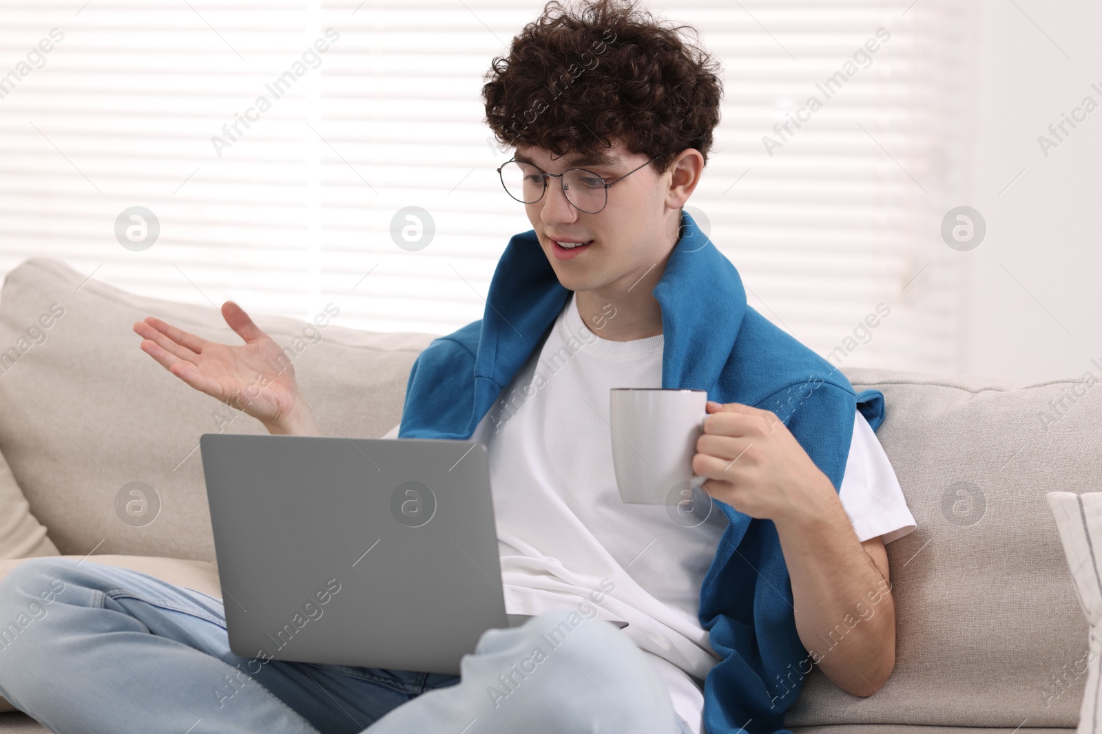 Photo of Teenager holding cup of drink working with laptop on sofa at home. Remote job