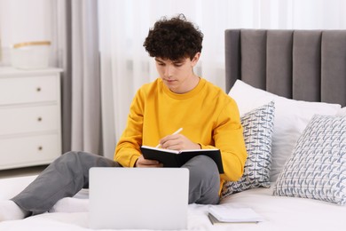 Photo of Teenager taking notes while working with laptop on bed at home. Remote job