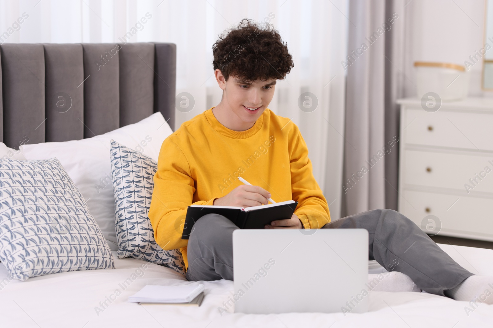 Photo of Teenager taking notes while working with laptop on bed at home. Remote job