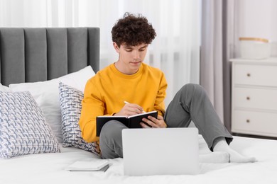 Photo of Teenager taking notes while working with laptop on bed at home. Remote job