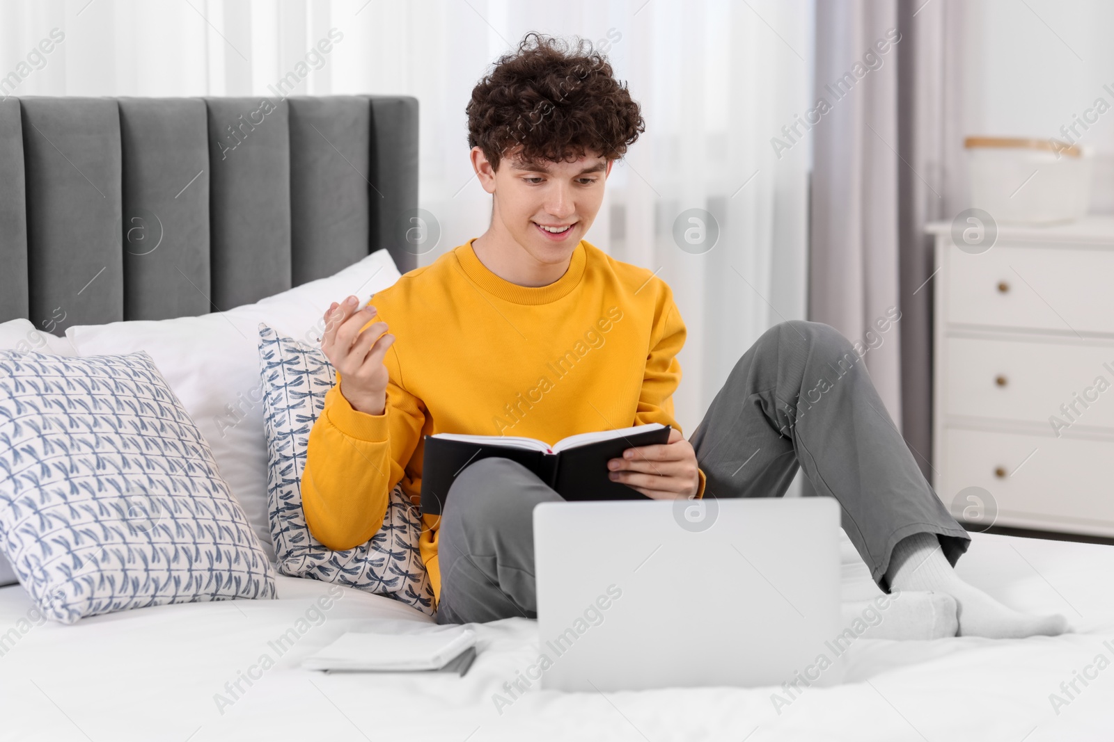 Photo of Teenager taking notes while working with laptop on bed at home. Remote job