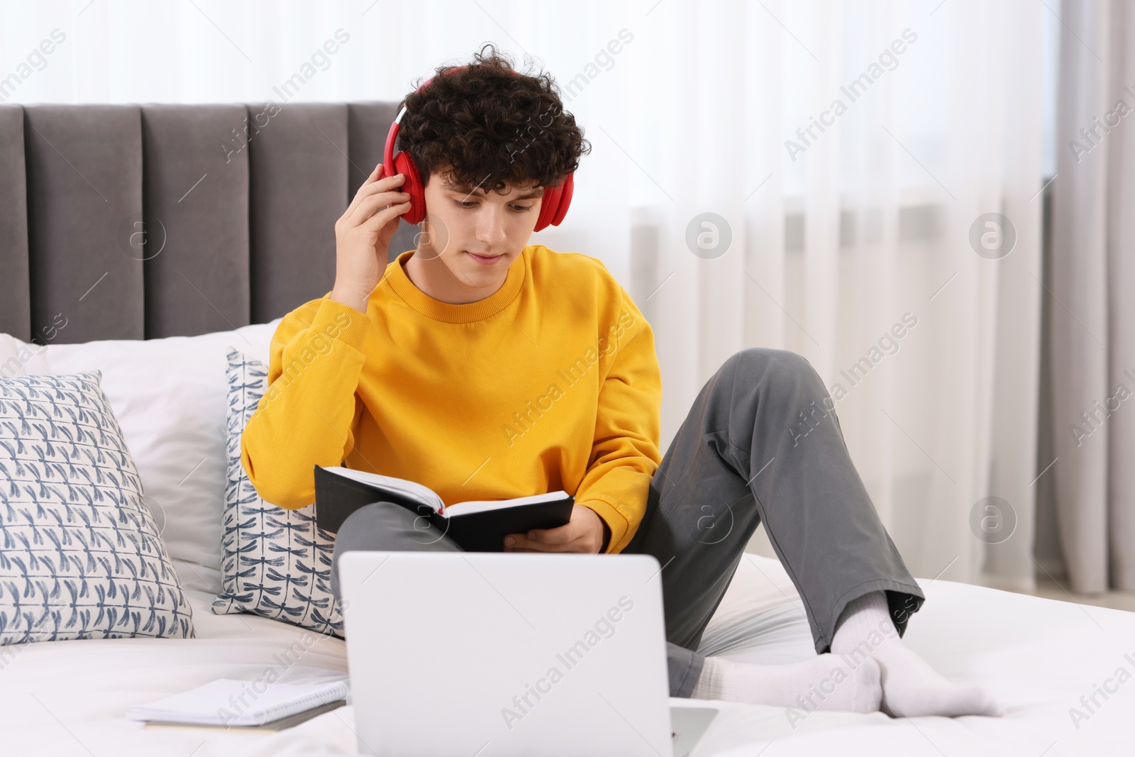 Photo of Teenager in headphones holding notebook and working with laptop on bed at home. Remote job