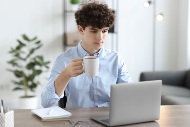 Teenager with cup of drink working on laptop at table indoors. Remote job