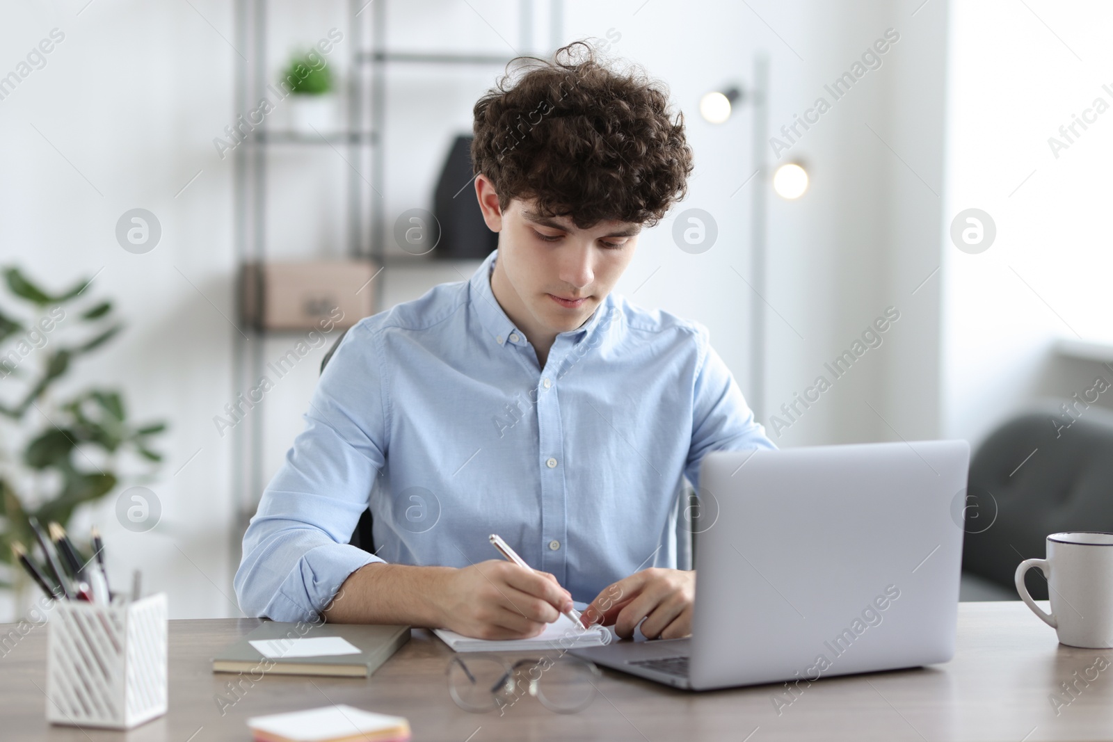 Photo of Teenager taking notes while working with laptop at table indoors. Remote job