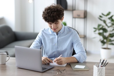 Teenager taking notes while working with laptop at table indoors. Remote job