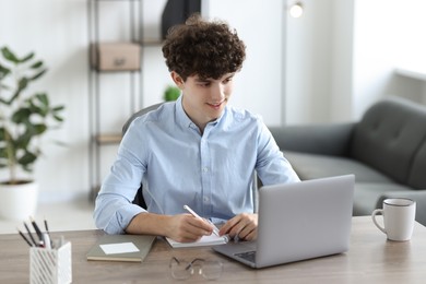 Teenager taking notes while working with laptop at table indoors. Remote job