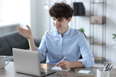 Photo of Teenager taking notes while working with laptop at table indoors. Remote job