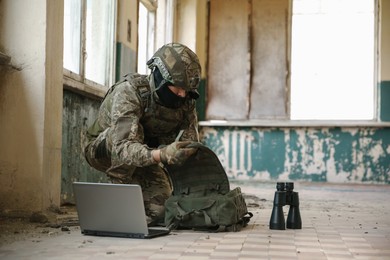 Photo of Military mission. Soldier in uniform using laptop and binoculars inside abandoned building