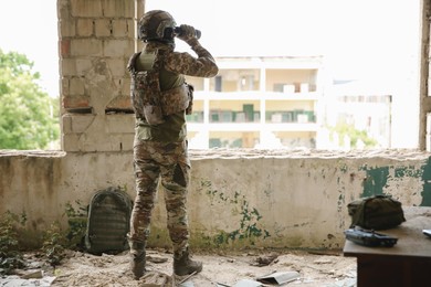 Photo of Military mission. Soldier in uniform with binoculars inside abandoned building, back view. Space for text