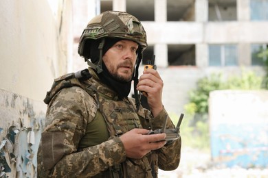 Photo of Military mission. Soldier in uniform with drone controller and radio transmitter near abandoned building outdoors