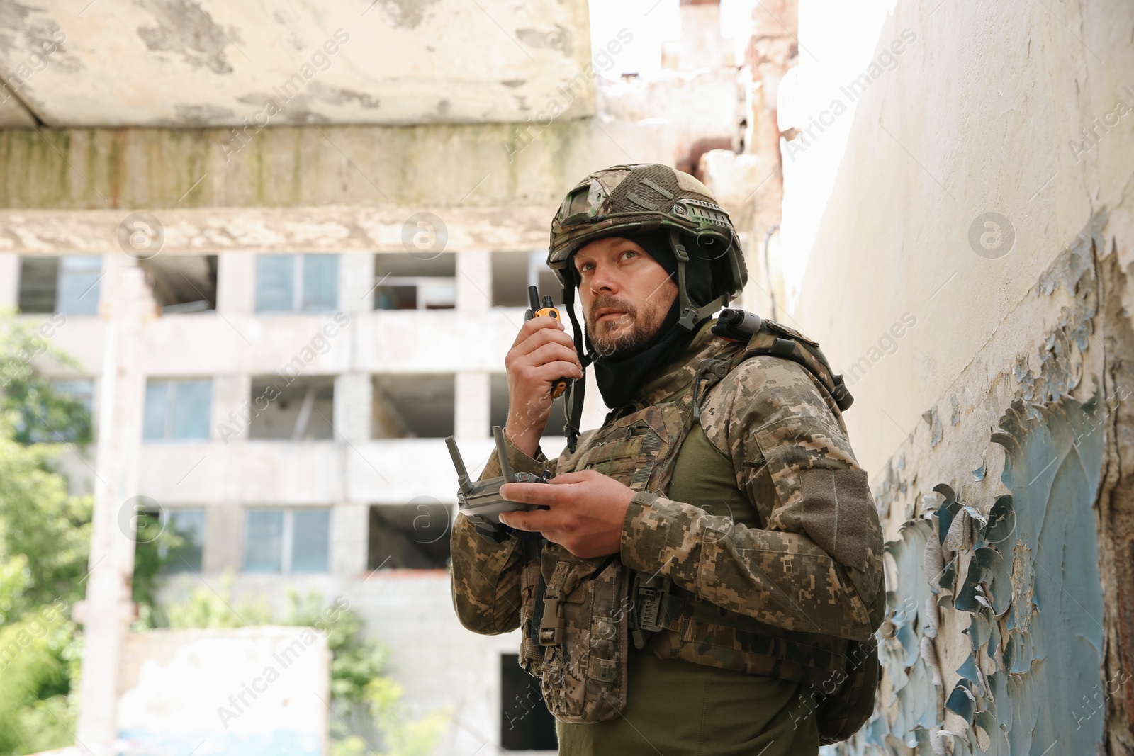 Photo of Military mission. Soldier in uniform with drone controller and radio transmitter near abandoned building outdoors, low angle view. Space for text
