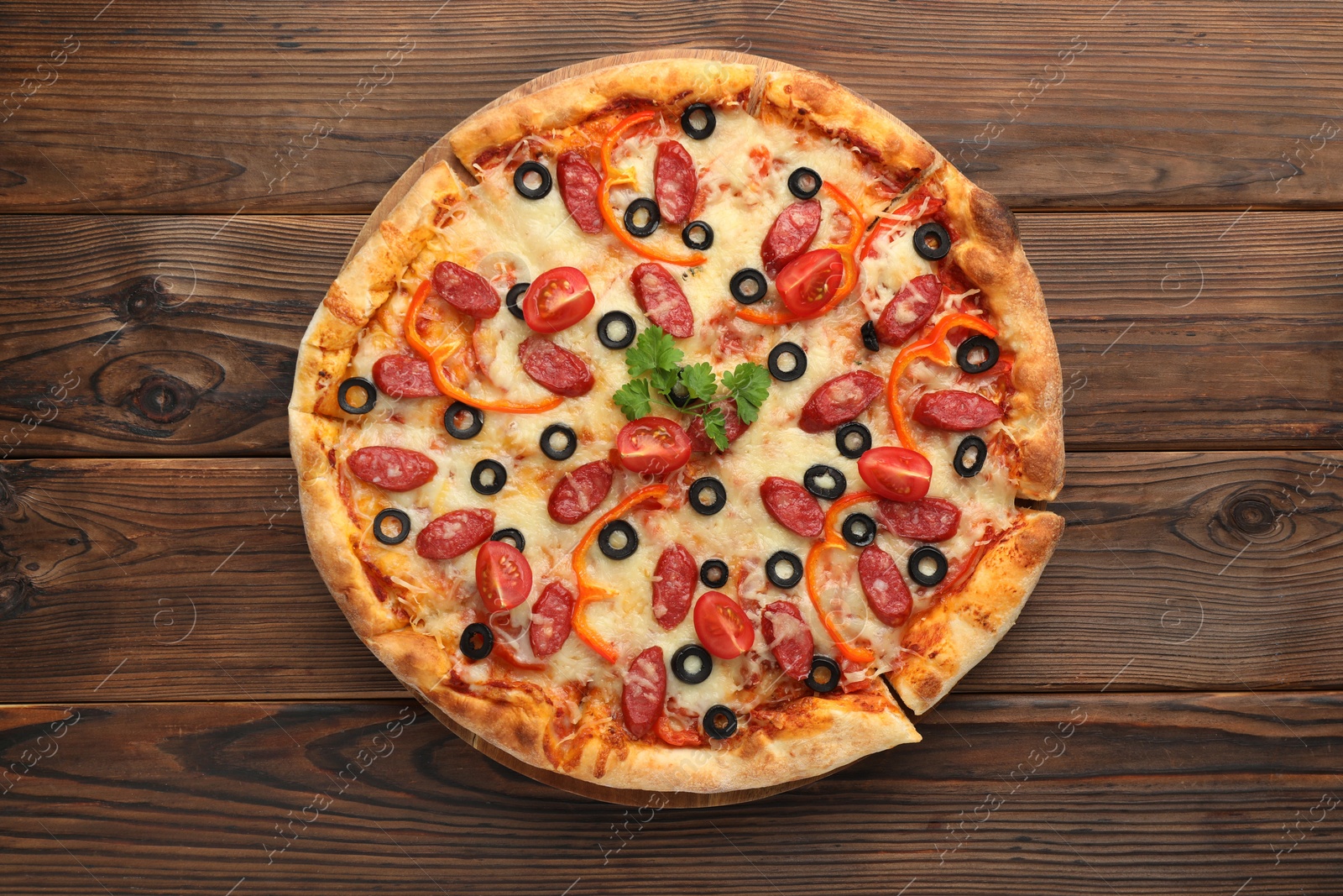 Photo of Tasty pizza with dry smoked sausages, tomato, olives, pepper and parsley on wooden table, top view